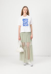 T-shirt blanc à manches courtes avec un motif floral bleu, associé à une jupe verte transparente à volants et des baskets blanches, portant un petit sac à main blanc.