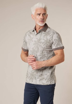 Homme plus âgé aux cheveux blancs portant un polo gris à manches courtes avec imprimé de palmiers et un pantalon bleu marine, debout les mains jointes.