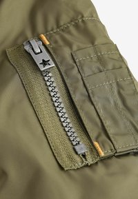 Tissu vert olive avec une fermeture éclair métallique munie d'une tirette en forme d'étoile, détails de poche cousus et accents de bord texturés.