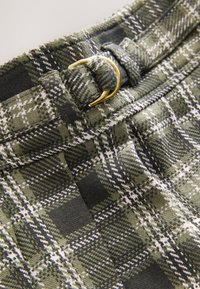 Green plaid fabric featuring a textured weave, a gold metal belt buckle, and a stitched waistband with contrasting black accents.
