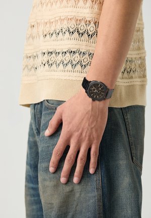 Polso maschile con orologio analogico nero, indossa un top beige lavorato a maglia e jeans blu, mano rilassata di lato contro uno sfondo semplice.