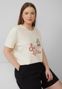 Cremfarbens Baumwoll-T-Shirt mit kurzen Ärmeln, das ein Grafikdesign einer Palme und den Text "Sea La Vie" in rosa und blauen Tönen zeigt. Schwarze Shorts.