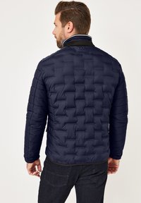 Mann in einer marineblauen gesteppte Jacke mit strukturierten Quadratmustern, steht mit dem Rücken zugewandt, kombiniert mit dunklen Jeans.