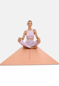 Yoga mat in zacht perzik met een centrale rode lijn voor uitlijning. Een persoon in lavendel sportkleding zit met gekruiste benen op de mat, op blote voeten.