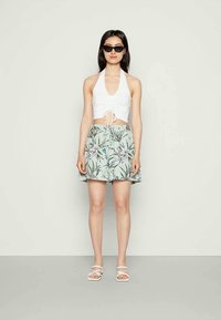 Mulher a usar óculos de sol pretos, top cropped branco com alças, calções florais verdes claros e sandálias brancas com tiras, em pé contra um fundo simples.