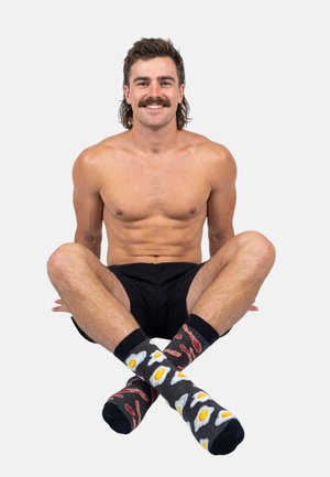 Uśmiechnięty mężczyzna bez koszuli z wąsami i mullet siedzi ze skrzyżowanymi nogami, ubrany w czarne szorty i niepasujące skarpetki z wzorami sadzonych jajek i bekonu.