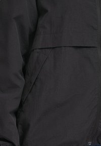 Gros plan d'une veste noire montrant une fermeture éclair à l'avant et une poche latérale inclinée avec des détails de couture sur le tissu.