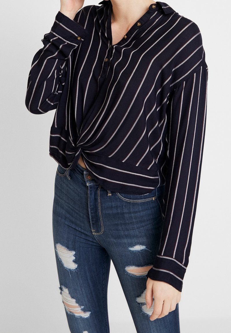 Femme portant une chemise bleu marine à manches longues avec des rayures blanches, boutonnée devant et ourlet torsadé, associée à un jean bleu déchiré.