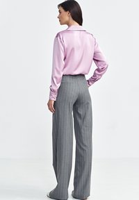 Różowa satynowa koszula z długim rękawem i kołnierzykiem, zestawiona z szarymi spodniami w szerokie paski. Gładka tekstura, dopasowany krój, widoczne detale guzików.