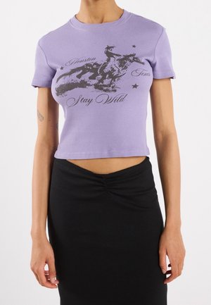 Kvinde iført en kort lavendel T-shirt med et sort grafisk motiv af en hesterytter og teksten "Stay Wild" kombineret med et sort skørt.