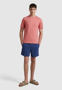 T-shirt rosso texture a maniche corte abbinato a pantaloni corti blu. L'outfit include sandali a scivolo verdi. Design semplice, senza pattern o loghi visibili.