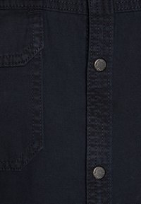 Chemise en denim foncé avec un devant à boutons, une poche poitrine simple et des boutons en métal, avec des coutures structurées et une finition en tissu texturé.