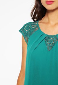 Turquoise blouse met kanten kapmouwtjes met cirkelpatronen, een subtiel plooitje bij de halslijn en een gladde stoffen textuur.