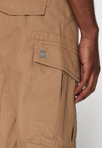 Close-up van een beige cargobroekzak met klep en metalen RAW-logo, gedragen door een persoon met een donkere huid en een ontspannen hand aan de zijkant.