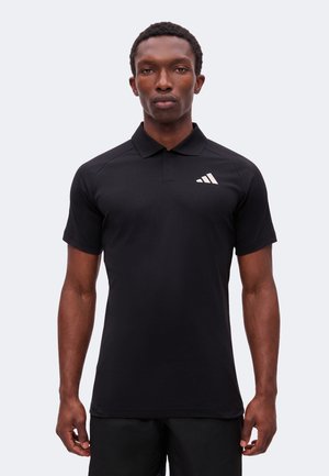 Homme portant un polo noir ajusté Adidas à manches courtes avec un logo blanc sur la poitrine gauche, debout devant un fond clair uni.