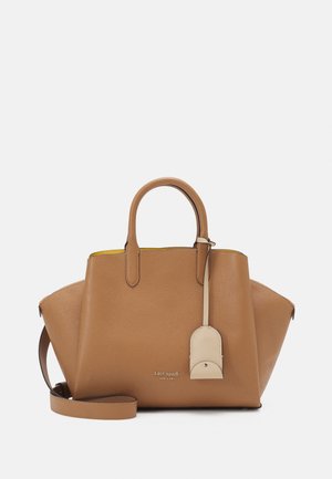 Sac à main en cuir tan Kate Spade avec poignées supérieures, bandoulière ajustable et étiquette beige suspendue sur fond blanc.