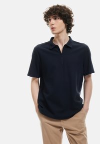 Marineblå kortærmet polo shirt med tekstureret stof, krave og lynlås halvvejs op. Bæres med lysebeige bukser, hænderne i lommerne.