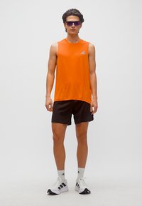 Νεαρός άνδρας που στέκεται φορώντας πορτοκαλί αμάνικη μπλούζα, μαύρα σορτς, λευκές κάλτσες και παπούτσια Adidas, και μοβ αθλητικά γυαλιά ηλίου.