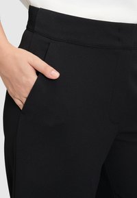 Hand in der Tasche von schwarzen, eng anliegenden Hosen mit Elastikbund, kombiniert mit einem weißen Oberteil.