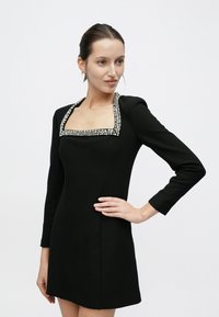 Μαύρο φόρεμα με μακριά μανίκια και τετράγωνη λαιμόκοψη, διακοσμημένο με περίτεχνη διάταξη χάντρων κατά μήκος της λαιμόκοψης. Εφαρμοστή γραμμή με μήκος μίνι.