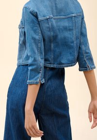 Henkilö, joka on pukeutunut lyhytvartiseen farkkutakkiin, jossa on kolmanneksen pituiset hihat, ja korkeavyötäröisiin farkkuihin, seisoo yksinkertaista taustaa vasten.