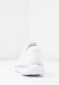 Vit träningsskor med en strukturerad mesh-övre och en metallisk silveraccent på hälen. Plan gummisula med en rundad design.