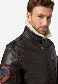 Braune Lederjacke mit Kunstfellkragen, Reißverschluss vorne und seitlichen Taschen. Verfügt über ein farbiges Patch am linken Ärmel.