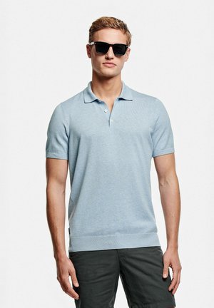 Jeune homme portant des lunettes de soleil noires, un polo à manches courtes bleu clair et un pantalon foncé, debout devant un fond blanc uni.