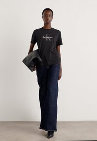 T-shirt noir en coton avec un logo Calvin Klein gris, associé à un jean évasé bleu marine et à une pochette noire. Bottes noires à talons épais.