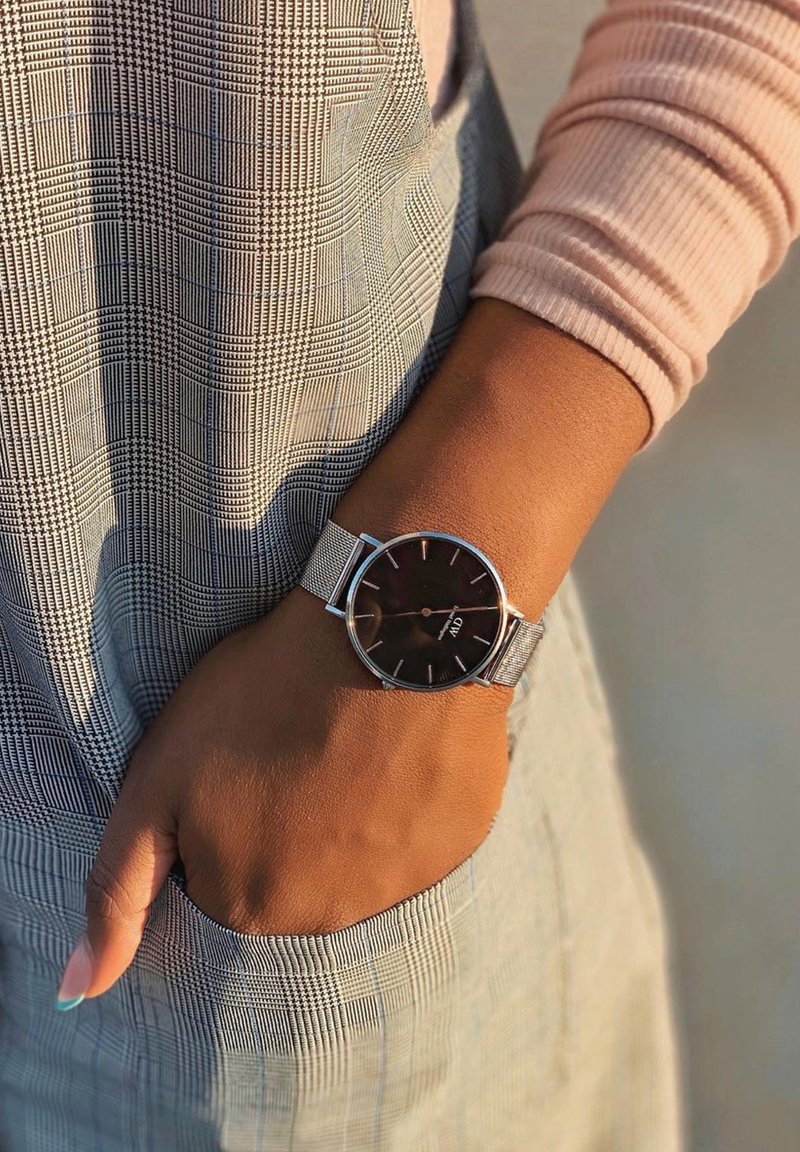 Poignet avec montre en maille argentée et cadran noir, reposant dans la poche d'un pantalon à carreaux gris, portant un haut rose clair à manches longues.