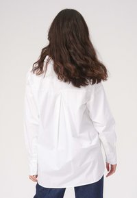 Hvid, langærmet skjorte med knaplukning og rygplyt-detalje, lige snit og knap-gemte manchetter, parret med mørkeblå jeans.