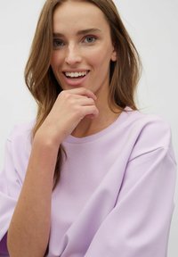 Svetle fialová mikina s kulatým výstřihem, volnějšími rukávy a měkkou texturou. Model se usmívá a opírá se o ruku.