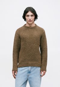 Persona joven con cabello oscuro hasta los hombros, vistiendo un suéter marrón texturizado y vaqueros de un azul claro, frente a un fondo blanco liso.