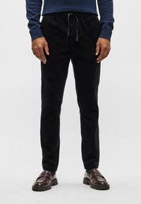 Calças de veludo cotelê pretas com uma silhueta ajustada, cintura elástica e detalhes de cordão. Combinadas com mocassins castanhos e um suéter azul.