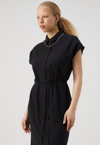 Rochie neagră cu butoni, cu mâneci scurte, dintr-un material texturat, cu talie accentuată printr-un brâu și cu guler. Însoțită de un colier din lanț auriu.