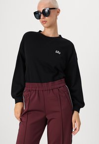 Crna dukserica s GAP logotipom, u kombinaciji s bordo trenirkom koja ima bočne pruge i elastičnu traku u struku. Model nosi velike sunčane naočale.