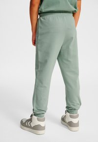 Persoon draagt saliegroene sweatpants en wit-grijze hoge sneakers, staand tegen een effen witte achtergrond.