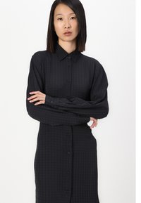 Robe chemise noire à boutons, avec un motif texturé, présentant des manches longues et un col, affichée sur un fond blanc uni.