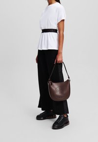 Hnědá kožená kabelka přes rameno s zakřiveným tvarem, perforovaným logem a pevným popruhem. Nositelná s bílým topem a širokými černými kalhotami.