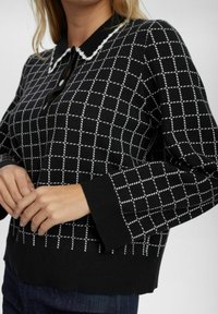 Pull en tricot noir avec un motif à carreaux blancs, un col, et des poignets côtelés. Boutons visibles au niveau du col.