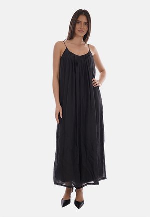 Mujer de pie con un vestido largo negro sin mangas con tirantes finos y tacones negros de punta afilada sobre un fondo blanco.