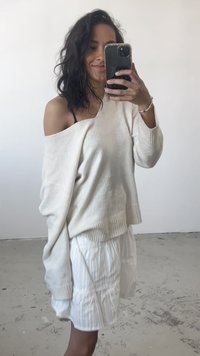 Suéter oversized em tom marfim com decote largo e bainha canelada, combinado com uma saia branca plissada. Textura suave, estilo casual.
