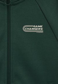 Temno zelena majica z zadrgo in belim napisom "GAME CHANGERS by BAD BOYS" na levem delu prsi, z ukrivljeno linijsko zasnovo.