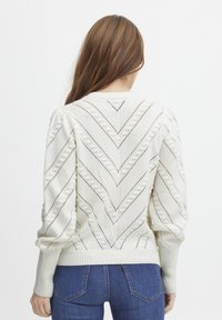 Pull en tricot blanc avec un motif en chevron, détails en dentelle et manches bouffantes ; poignets et ourlet côtelés, assorti à un jean bleu.