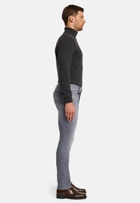 Maglione a collo alto di colore grigio scuro, aderente, con maniche lunghe; abbinato a jeans skinny grigio chiaro e scarpe eleganti marroni lucide. Vista laterale.