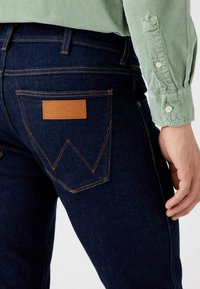 Jeans de mezclilla de un azul oscuro con un parche de cuero de Wrangler en el bolsillo trasero, con costuras naranjas y un diseño de pierna recta.