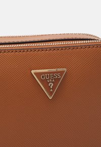 Cartera sintética marrón texturizada con cremallera plateada y una placa triangular del logo de GUESS USA en la parte frontal.