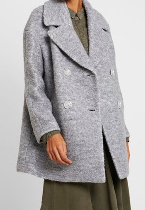 Personne portant un manteau en laine texturé gris clair croisé sur une chemise boutonnée et une jupe vert olive.