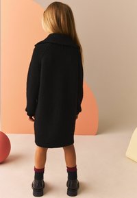 Abito nero in maglia con un grande colletto, maniche lunghe e texture a coste. Abbinato a stivali neri e calze bordeaux. Lo sfondo presenta forme morbide.