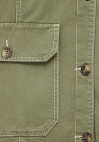 Olivengrønt denimstof med kontrasterende hvid stikning, en lommepatch og en afrundet knapdetalje på siden.